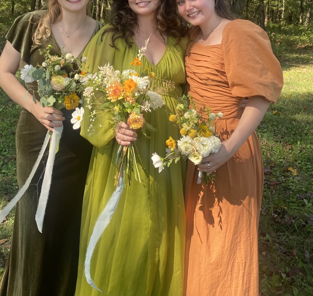 Three friends in green and orange dresses holding wildflower bouquets