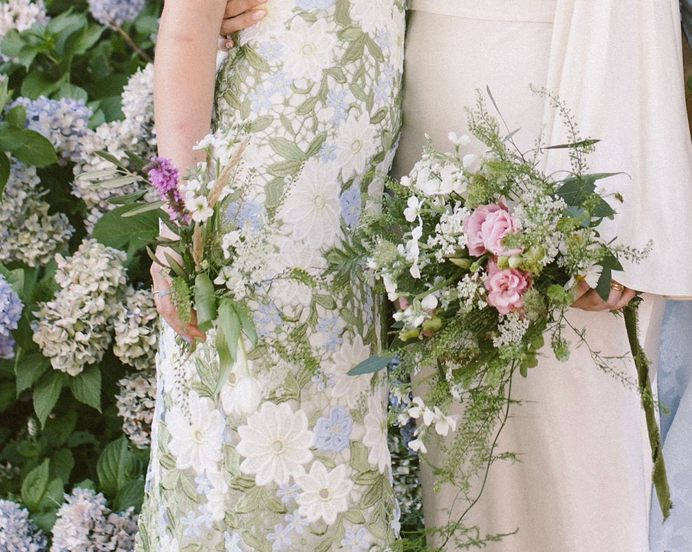 Two bridesmaids holding wildflower bouquets beside a hydrangea bush