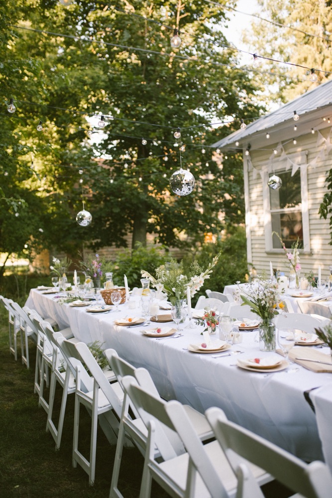 Two long tables in a garden under string lights and disco balls, set with wildflowers and white folding chairs