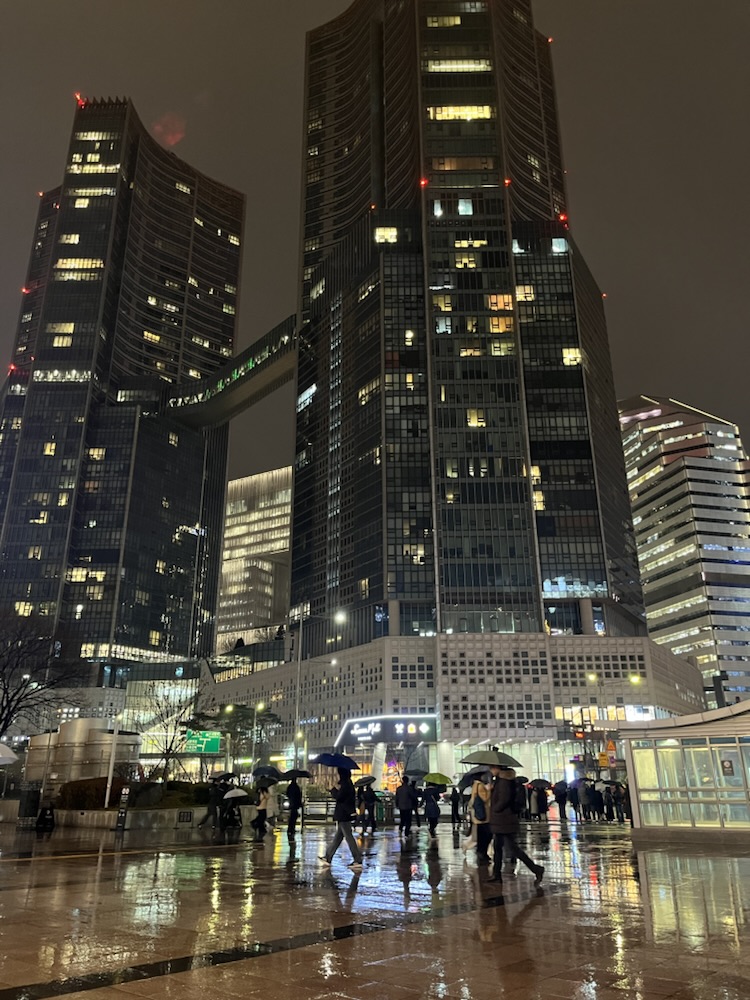 Umbrellas crossing a rain-slick plaza under lit towers, Seoul